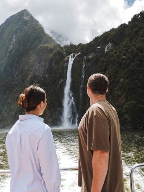 Couple on Milford Sound cruise viewing waterfall and lush mountains.