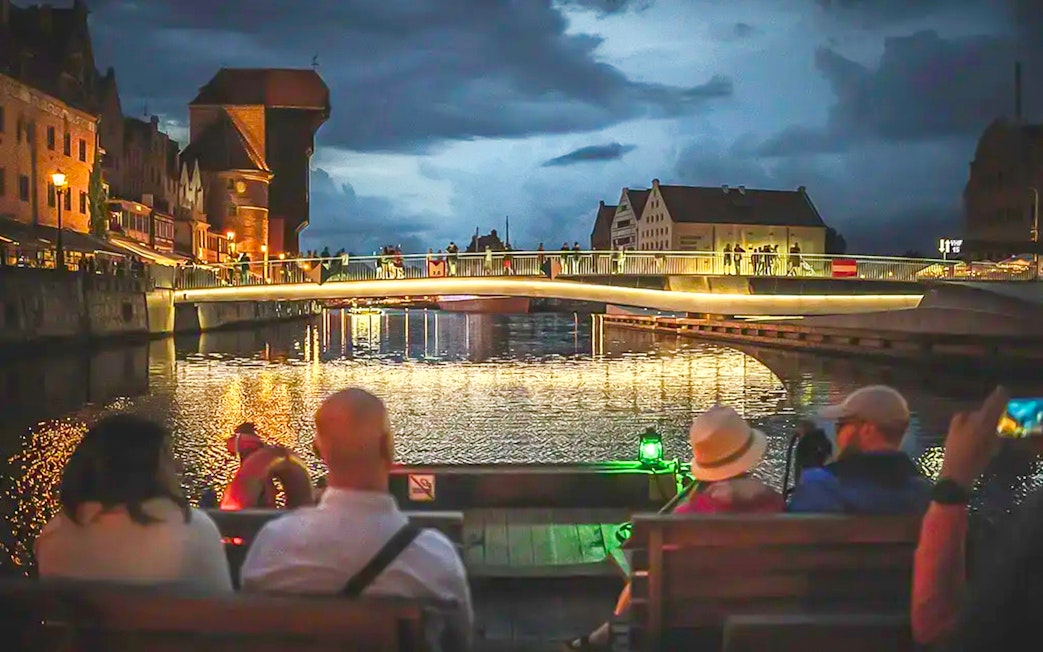 Guests on a historical boat during sunset river cruise in Gdansk, viewing illuminated bridge.