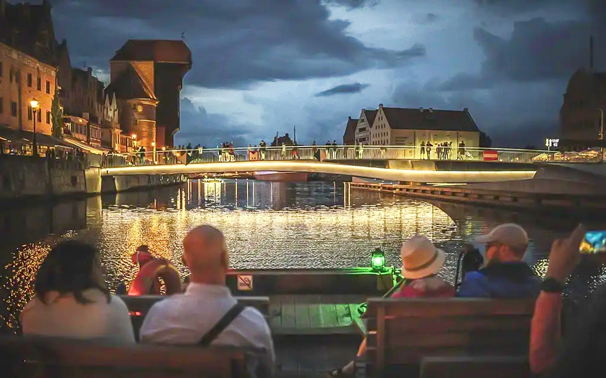 Guests on a historical boat during sunset river cruise in Gdansk, viewing illuminated bridge.