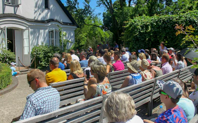 Visitors seated outside Fryderyk Chopin's birthplace in Poland, surrounded by greenery.