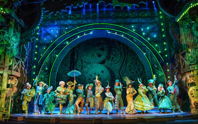 Cast of Wicked performing on stage with elaborate costumes and green-lit gears in the background.