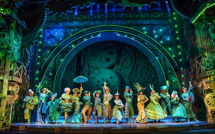 Cast of Wicked performing on stage with elaborate costumes and green-lit gears in the background.