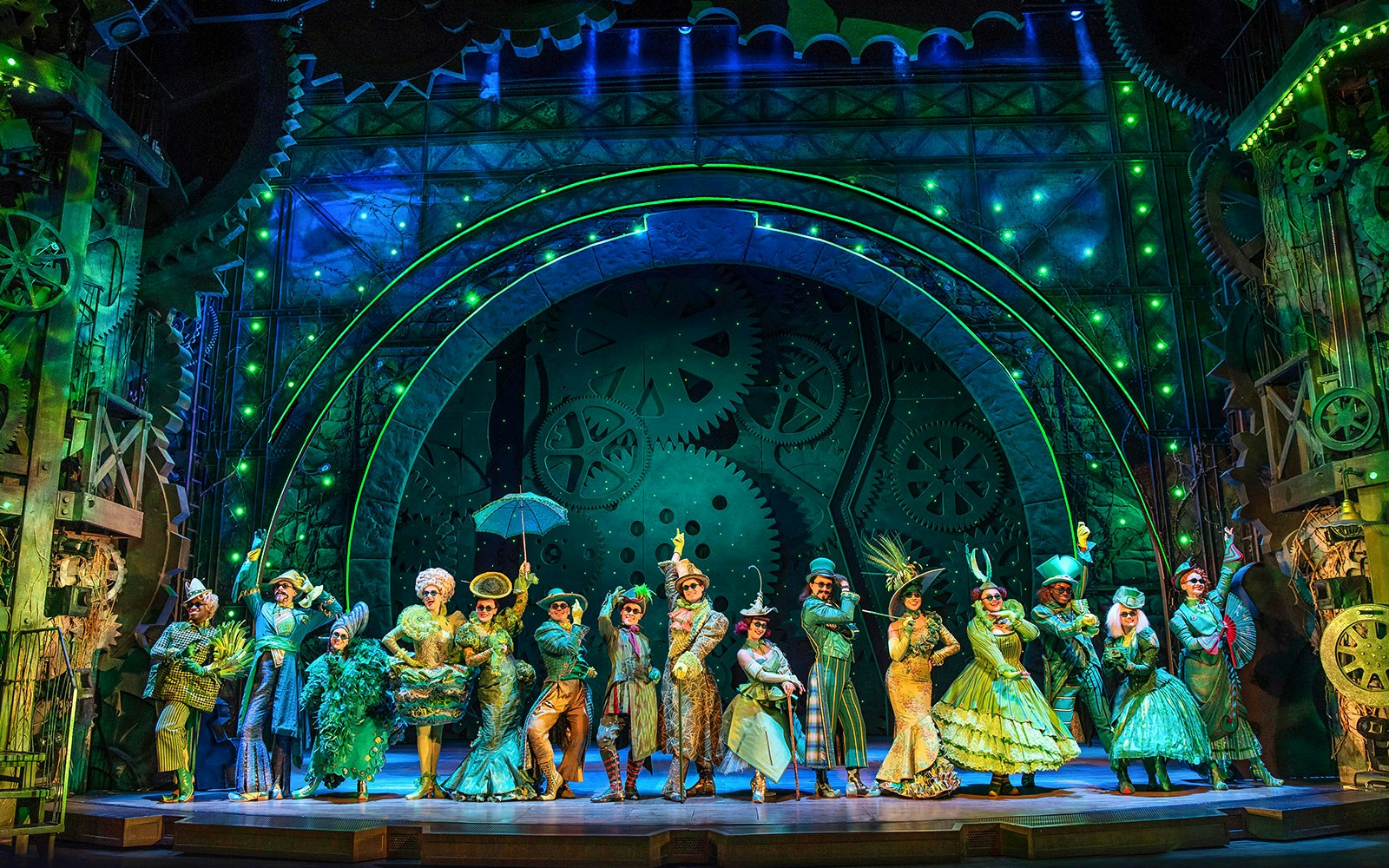 Cast of Wicked performing on stage with elaborate costumes and green-lit gears in the background.