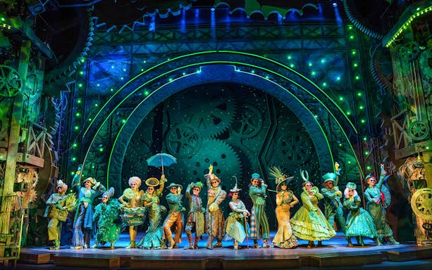 Cast of Wicked performing on stage with elaborate costumes and green-lit gears in the background.