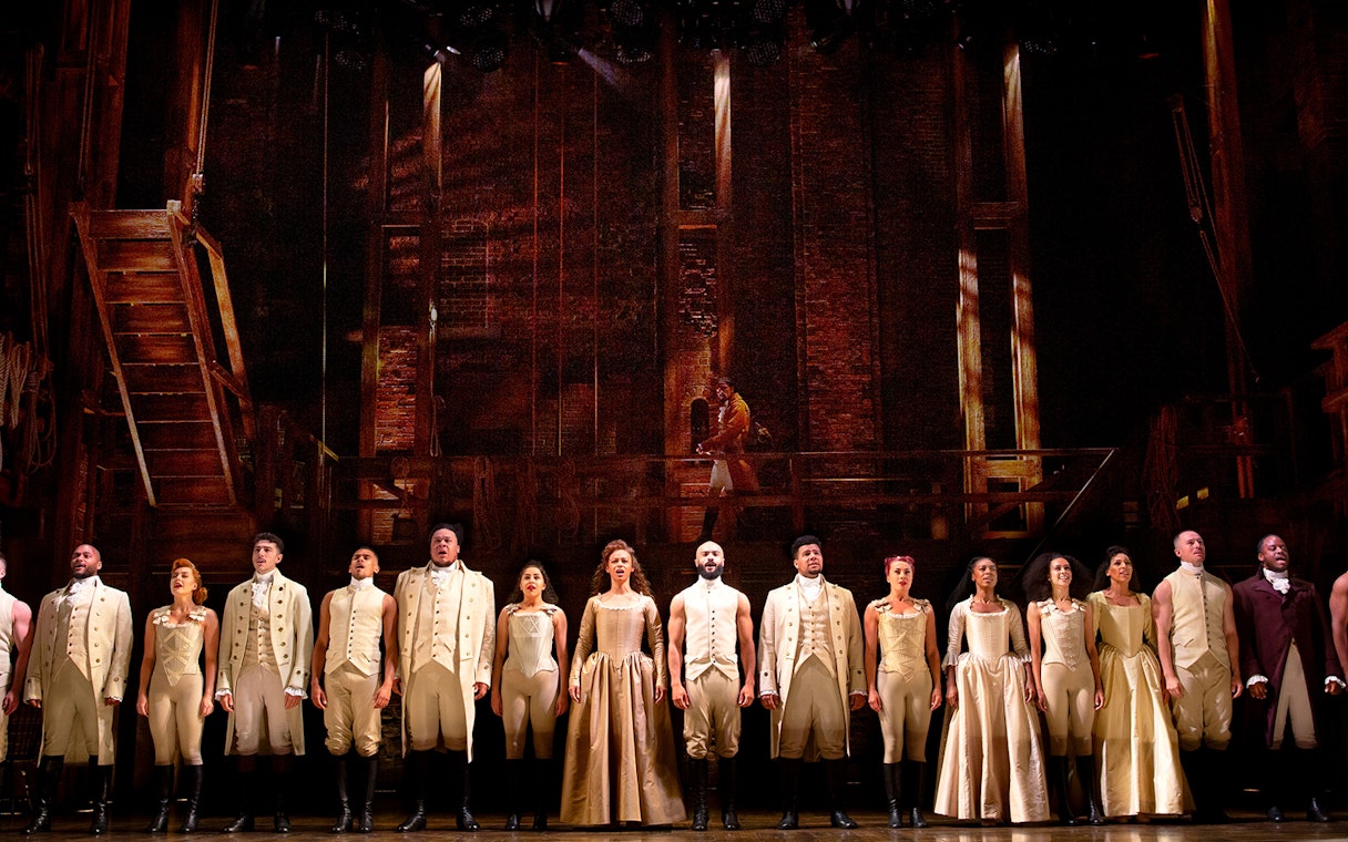 Cast of Hamilton performing on stage in period costumes.
