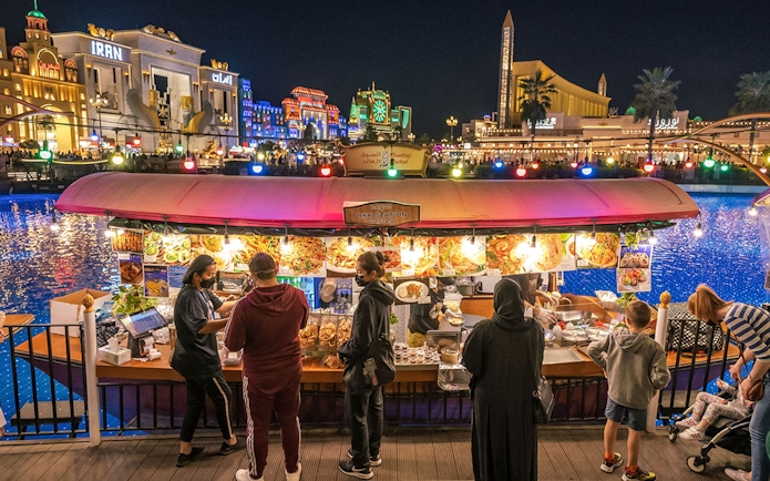 Floating market stall with visitors at Global Village Dubai, illuminated buildings in the background.