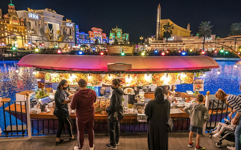 Floating market stall with visitors at Global Village Dubai, illuminated buildings in the background.
