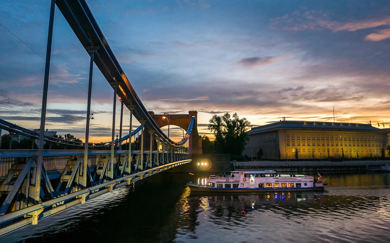 Boat cruising on the Vistula River at sunset near a lit-up bridge and building.