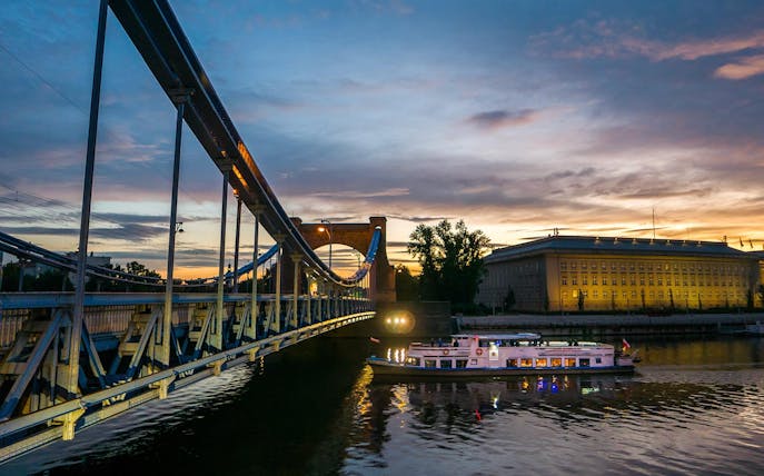 Boat cruising on the Vistula River at sunset near a lit-up bridge and building.