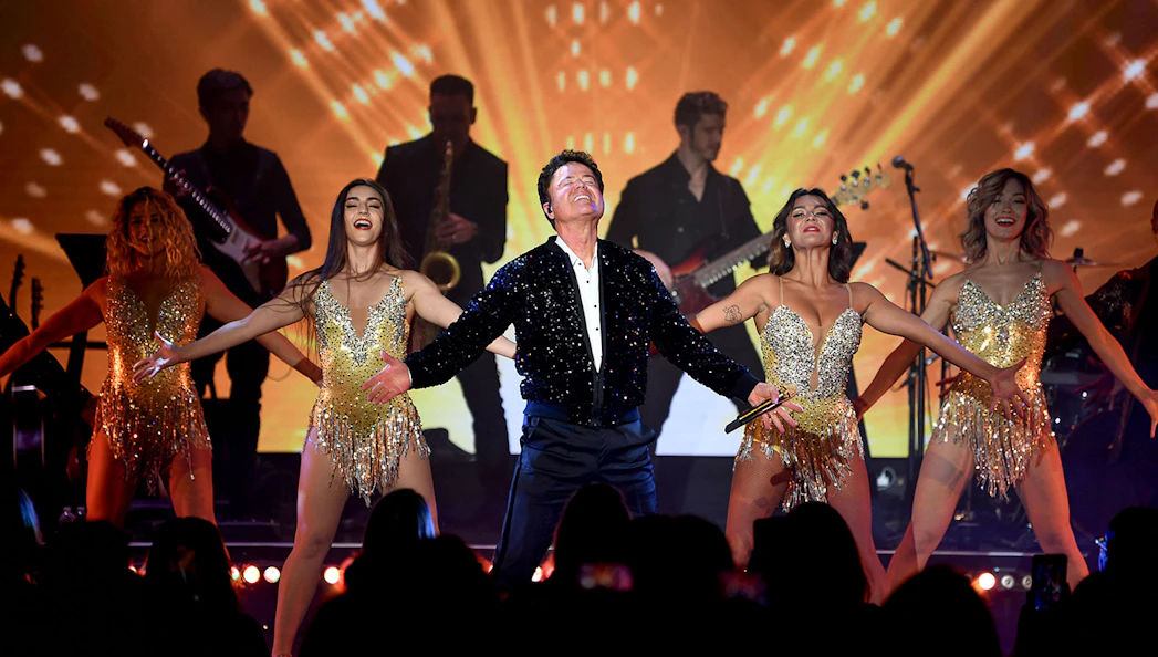 Performer on stage with dancers and band, vibrant light show in background.