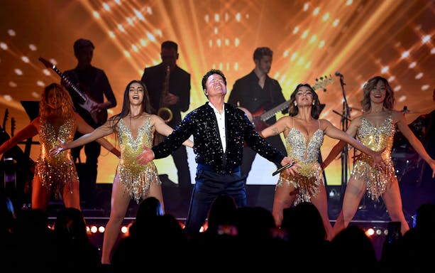 Performer on stage with dancers and band, vibrant light show in background.