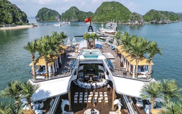 Aerial view of cruise deck with pool and palm trees, surrounded by limestone islands in Halong Bay.