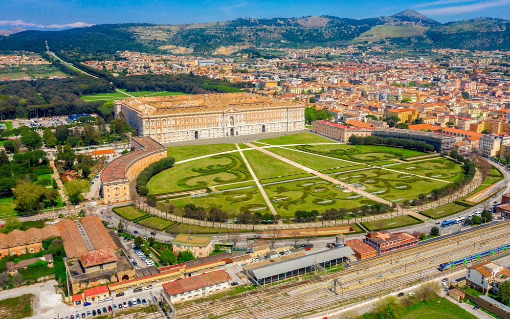 Aerial view of the Royal Palace of Caserta in Italy with expansive gardens and surrounding cityscape.