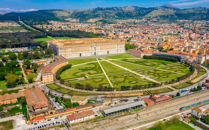 Aerial view of the Royal Palace of Caserta in Italy with expansive gardens and surrounding cityscape.
