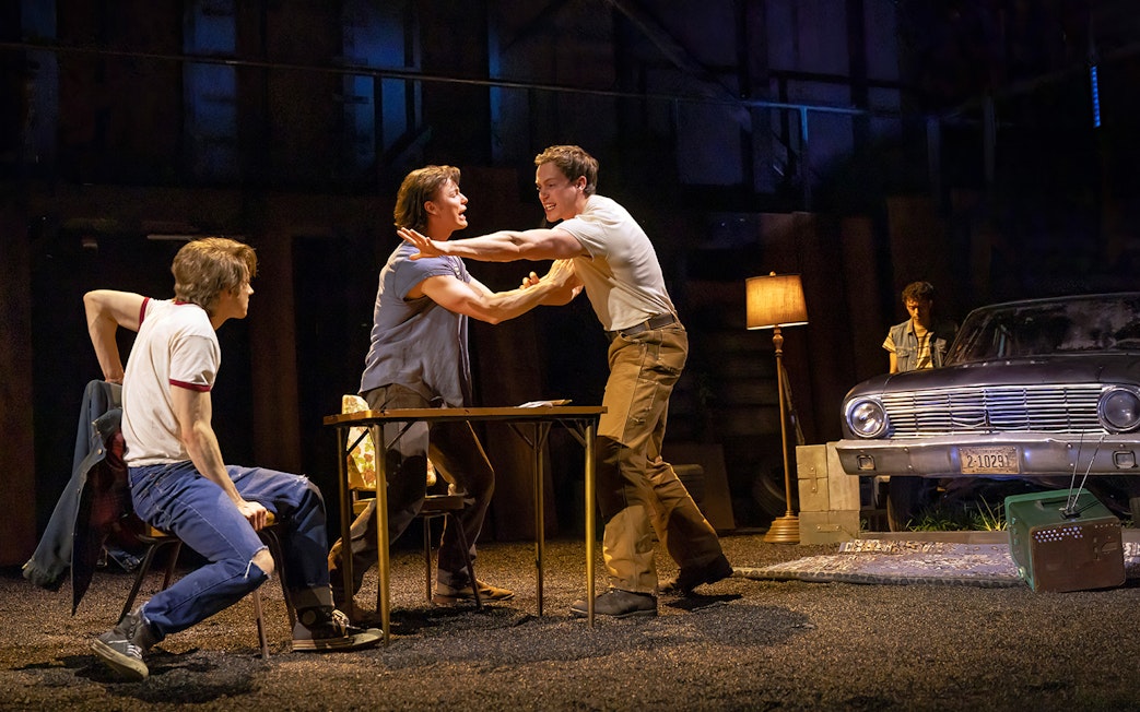Men in a tense scene from "The Outsiders" stage play with a vintage car in the background.