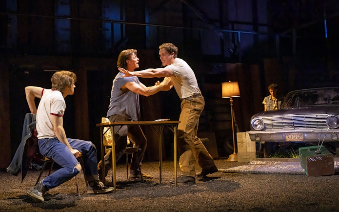 Men in a tense scene from "The Outsiders" stage play with a vintage car in the background.
