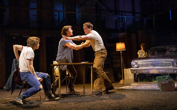 Men in a tense scene from "The Outsiders" stage play with a vintage car in the background.