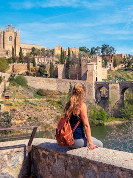 Woman sitting by the Tagus River with view of Toledo's medieval walls, Spain.