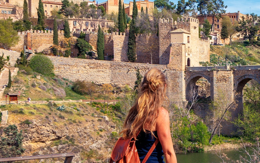 Woman sitting by the Tagus River with view of Toledo's medieval walls, Spain.