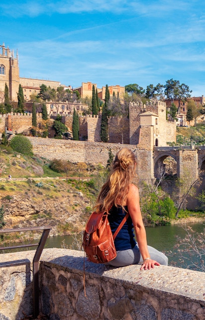Woman sitting by the Tagus River with view of Toledo's medieval walls, Spain.