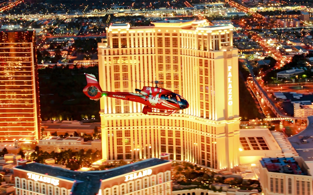 Red helicopter flying over Las Vegas Strip at night with Palazzo Hotel and Treasure Island in view.