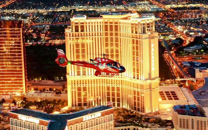 Red helicopter flying over Las Vegas Strip at night with Palazzo Hotel and Treasure Island in view.
