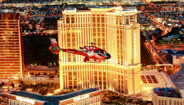 Red helicopter flying over Las Vegas Strip at night with Palazzo Hotel and Treasure Island in view.