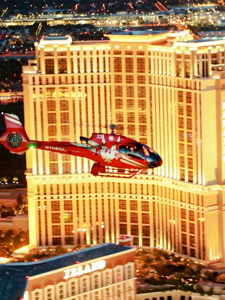 Red helicopter flying over Las Vegas Strip at night with Palazzo Hotel and Treasure Island in view.