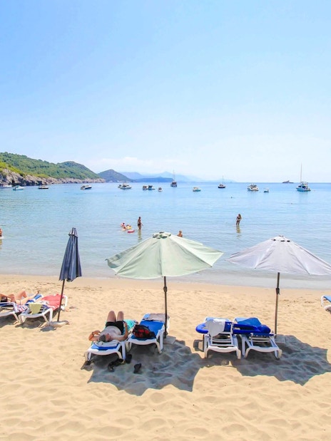 Sunbathers and umbrellas on Šunj Beach with boats in the bay, Lopud Island, Croatia.