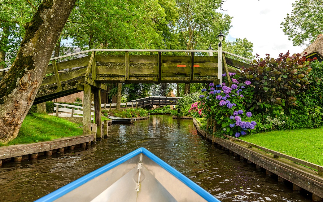 Boat navigating canal under wooden bridge in Giethoorn, Netherlands, with vibrant flowers and greenery.