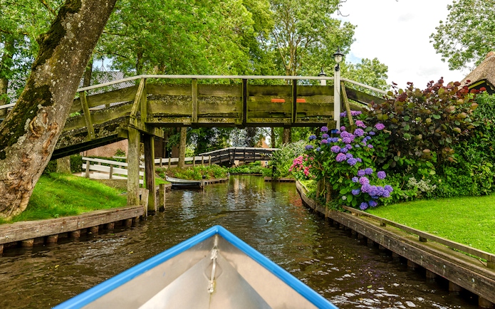 Boat navigating canal under wooden bridge in Giethoorn, Netherlands, with vibrant flowers and greenery.