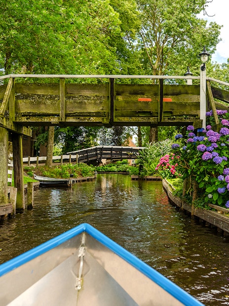 Boat navigating canal under wooden bridge in Giethoorn, Netherlands, with vibrant flowers and greenery.