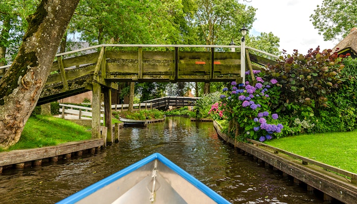 Boat navigating canal under wooden bridge in Giethoorn, Netherlands, with vibrant flowers and greenery.