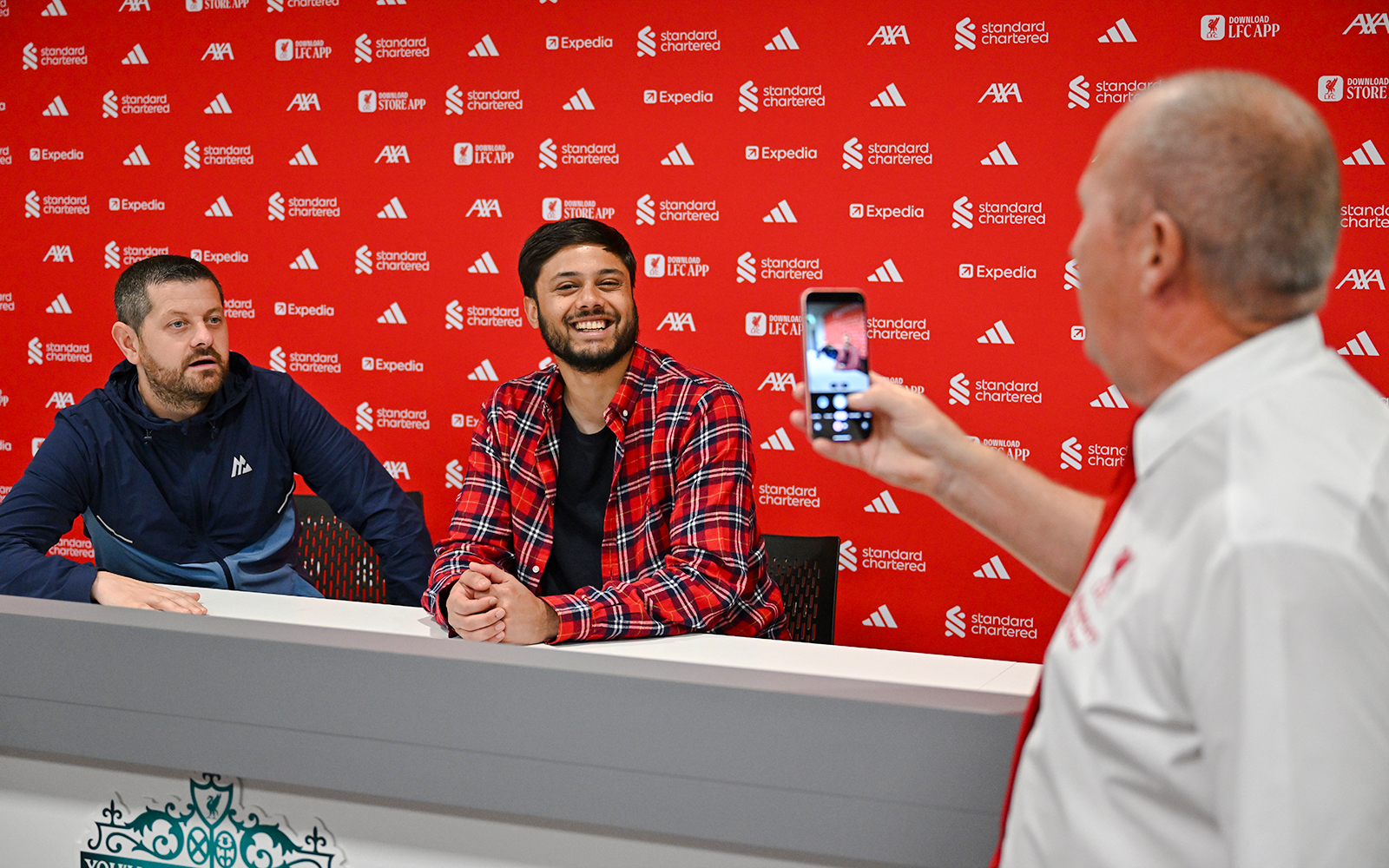 Guests in the press room at Anfield, with one taking a photo of two others at a table.
