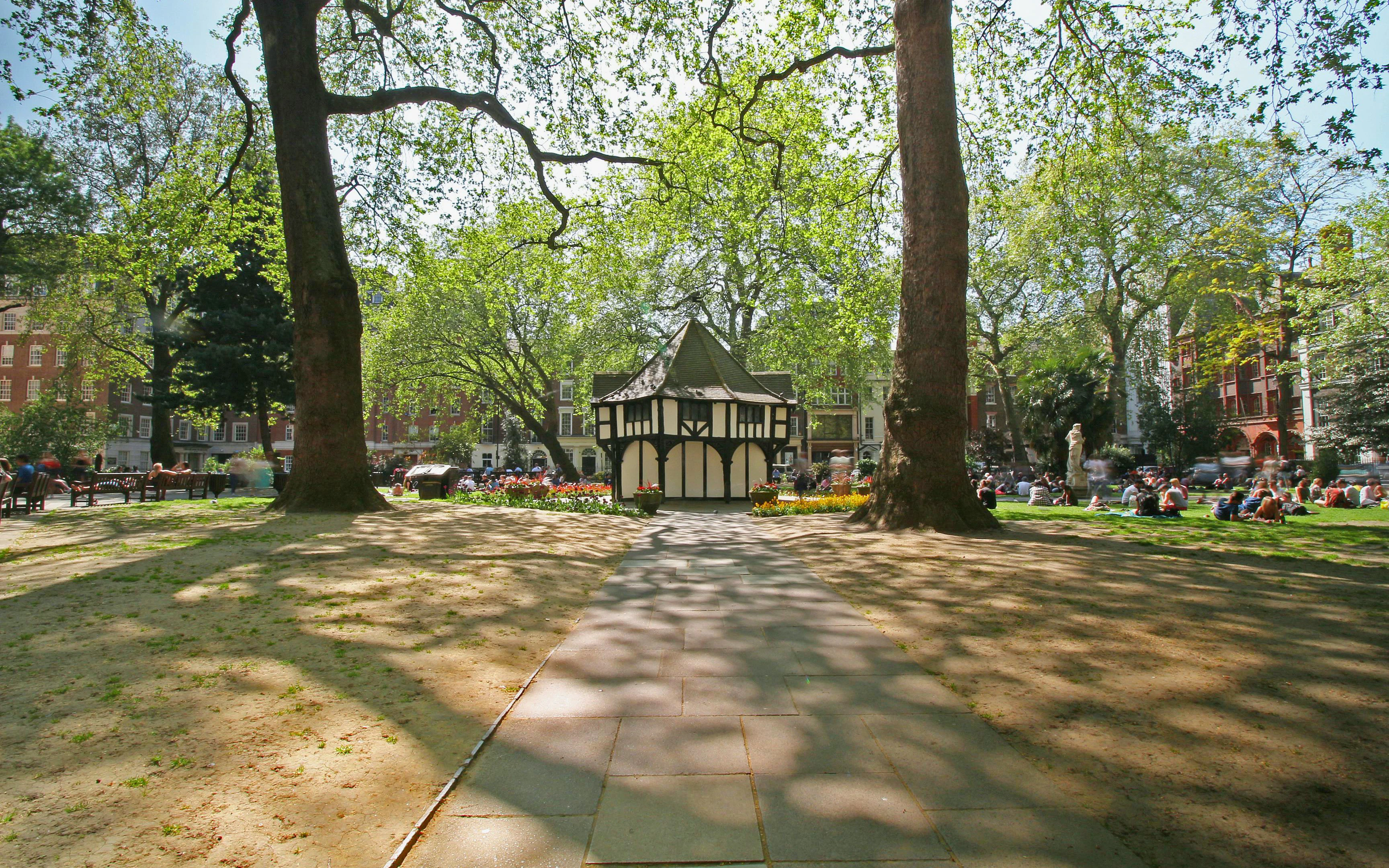 Pathway leading to a pavilion in Soho Square, London, surrounded by trees and people relaxing.