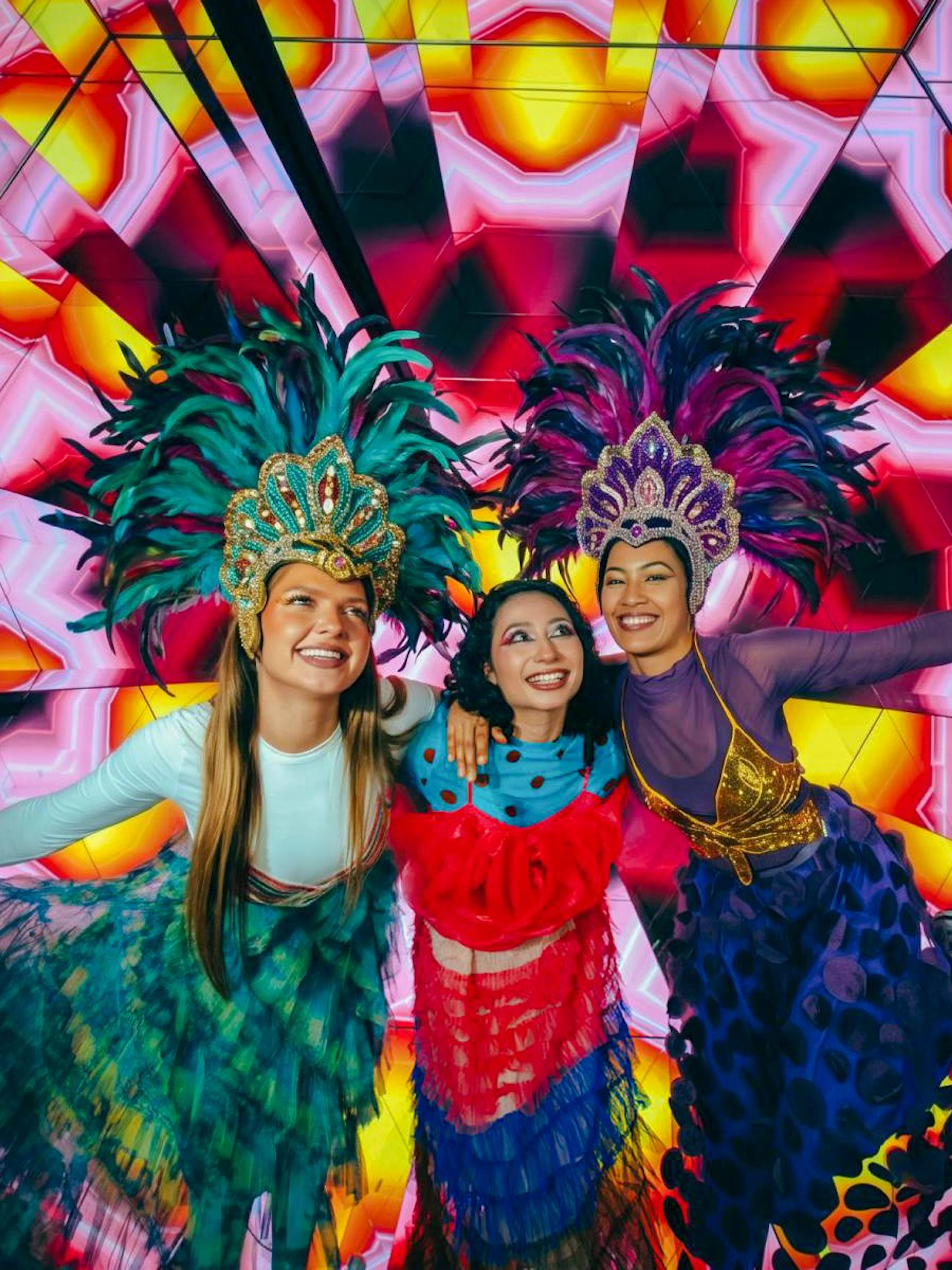 Three people in vibrant costumes and feathered headdresses at Electric Carnival, House of Hype.