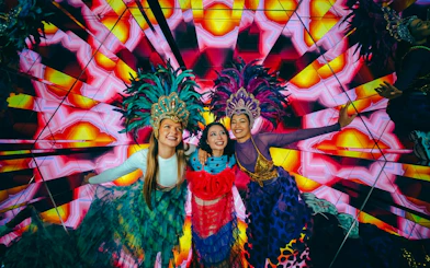 Three people in vibrant costumes and feathered headdresses at Electric Carnival, House of Hype.