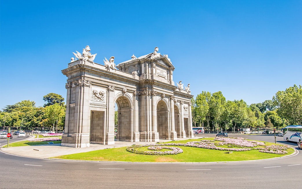Puerta de Alcalá in Madrid, Spain, surrounded by trees and flowers on a sunny day.