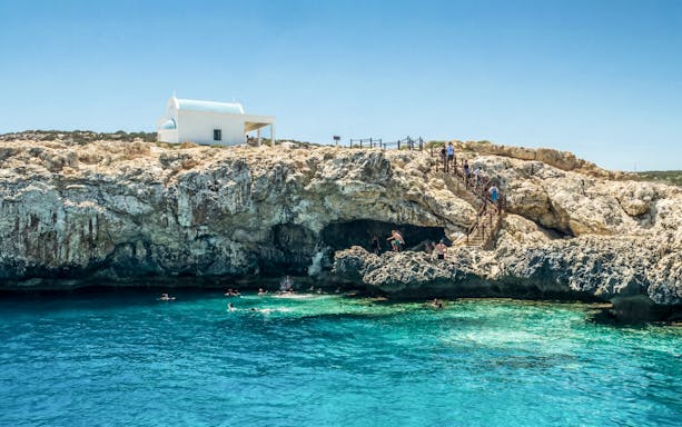 Swimmers explore Blue Lagoon caves near Protaras with a white chapel on the cliff.
