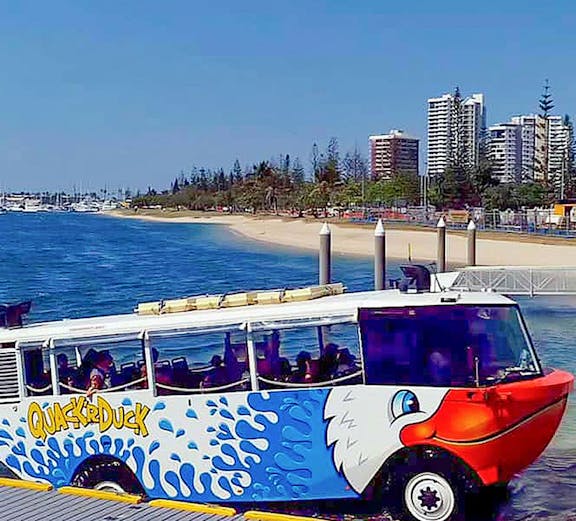 Quack'R Duck amphibious vehicle on a beachside dock with city skyline in the background.