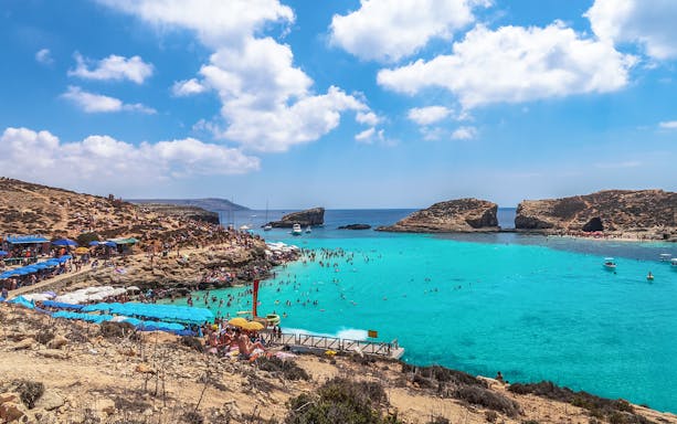 Blue Lagoon in Malta with tourists swimming and sunbathing, surrounded by rocky cliffs and clear blue water.