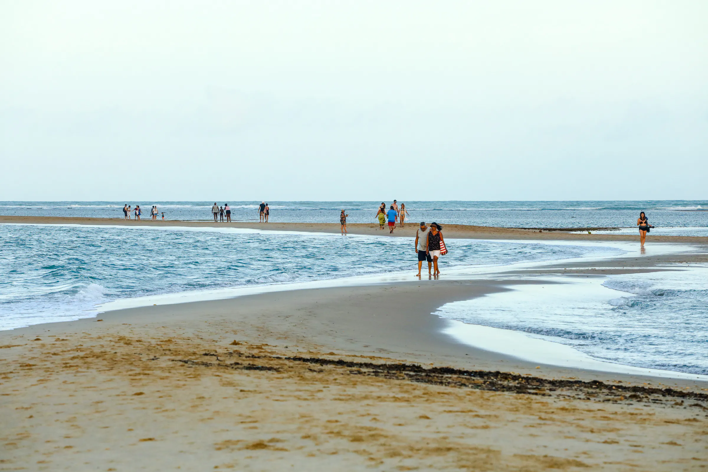 People walking on a sandy beach at Coroa Vermelha, Porto Seguro, with ocean waves in the background.