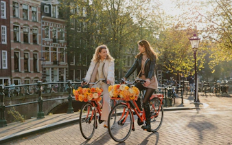 Two people biking along a canal in Amsterdam with flower baskets.