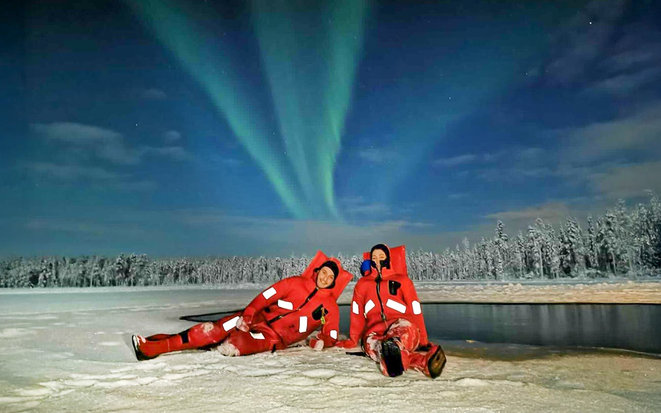 Guests in red suits relaxing on a frozen Lapland lake under Northern Lights.