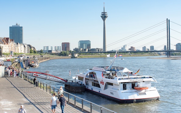 Panorama cruise boat docked on the Rhine River with Düsseldorf skyline and Rheinturm tower.