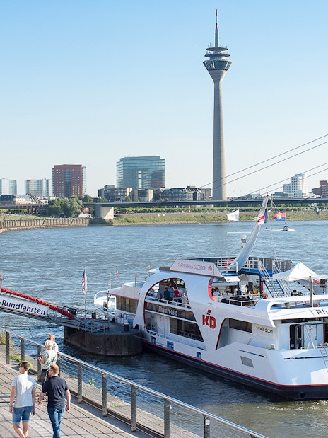 Panorama cruise boat docked on the Rhine River with Düsseldorf skyline and Rheinturm tower.