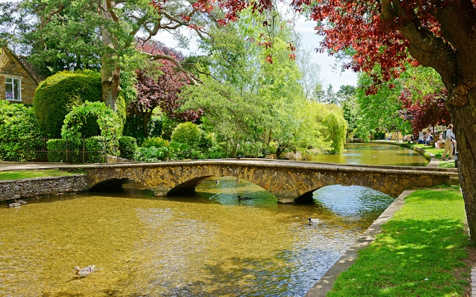 Stone bridge over a tranquil river in Bourton-on-the-Water, England, surrounded by lush greenery.