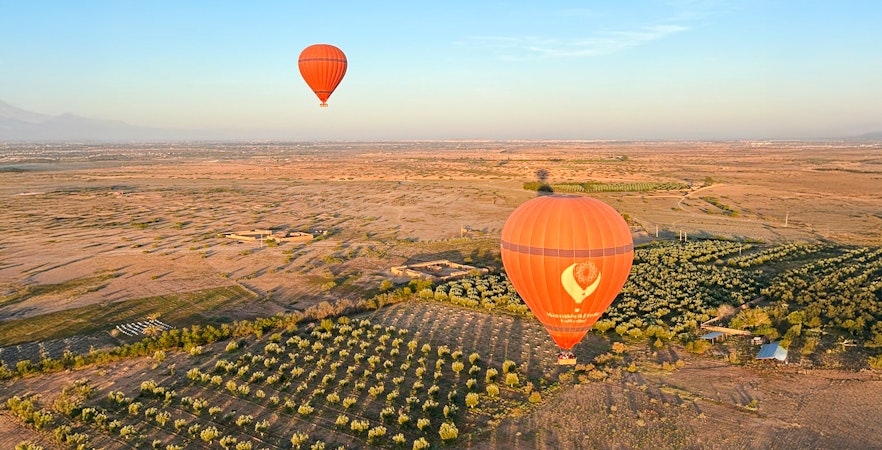 Hot air balloons floating over Marrakech landscape with fields and distant mountains.