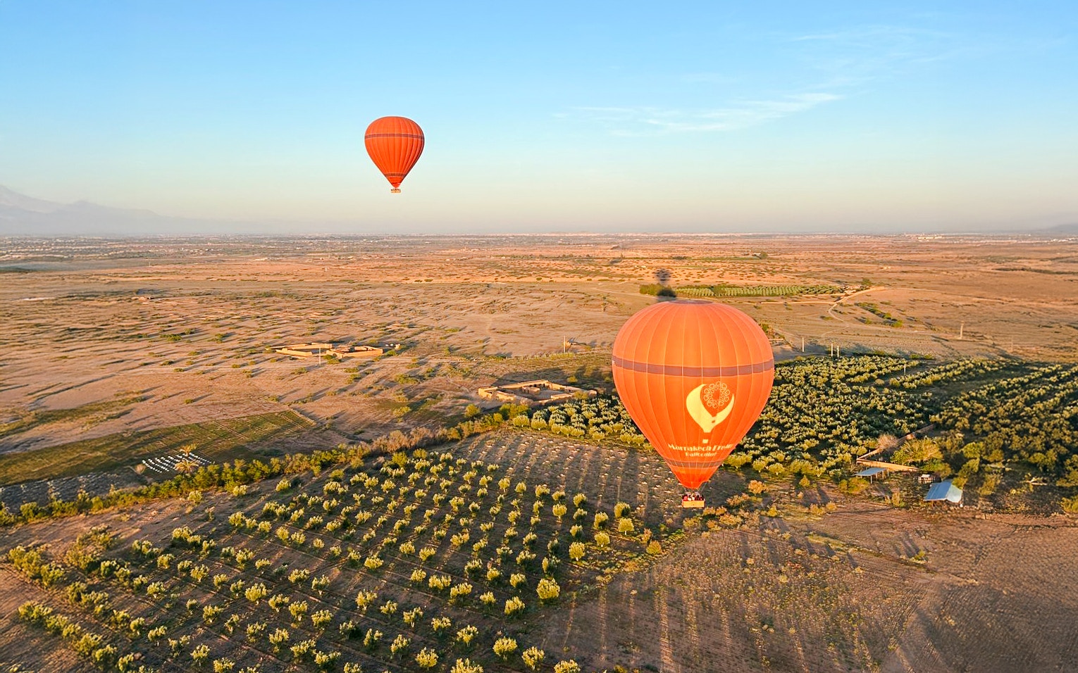 Hot air balloons floating over Marrakech landscape with fields and distant mountains.