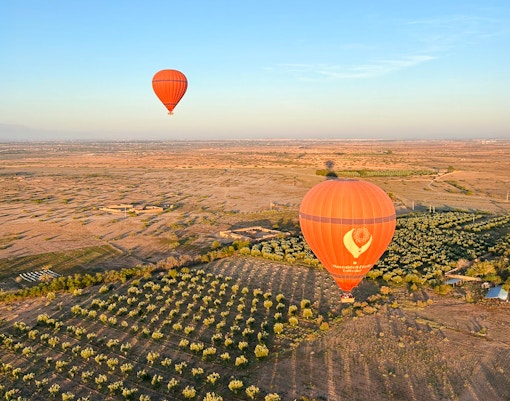 Hot air balloons floating over Marrakech landscape with fields and distant mountains.